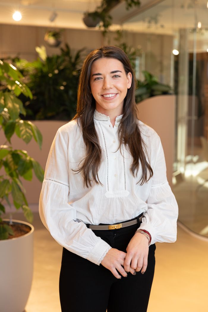 Vrouw zit binnen op een lichte kantoorlocatie en glimlacht richting de camera, met planten op de achtergrond.