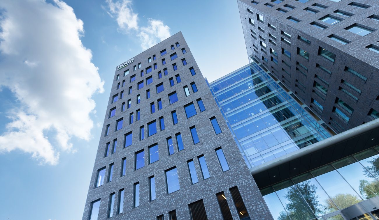 Grijze baksteentorens met glazen atrium en slanke raamstroken, tegen blauwe lucht — Ontwikkeling Dorgelo Building Eindhoven