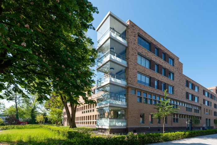 Modern woongebouw met bakstenen gevel en glazen balkons omringd door groen en bomen op een zonnige dag.
