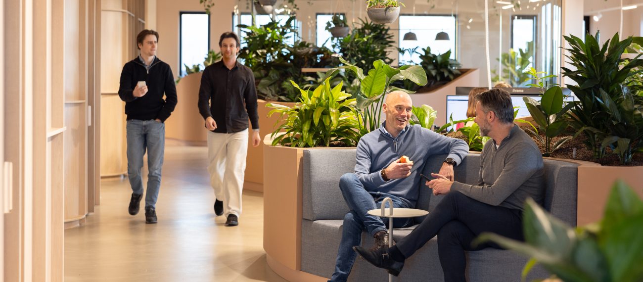Twee mannen zitten pratend op een bankje in een modern kantoor met veel planten, terwijl twee andere mannen langslopen op de achtergrond