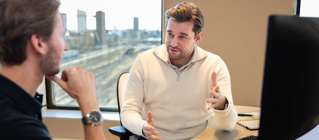 Twee medewerkers voeren een gesprek aan een bureau in een kantoor met uitzicht op spoorrails; de persoon rechts spreekt en gebaart terwijl de ander luistert.