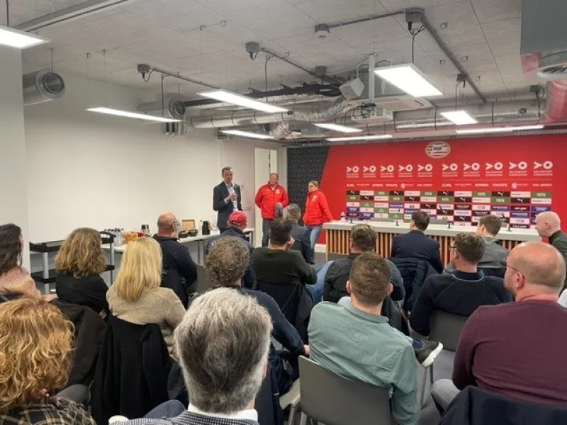 Presentatie voor zittend publiek in ruimte met sponsorwand en zichtbare installaties aan plafond