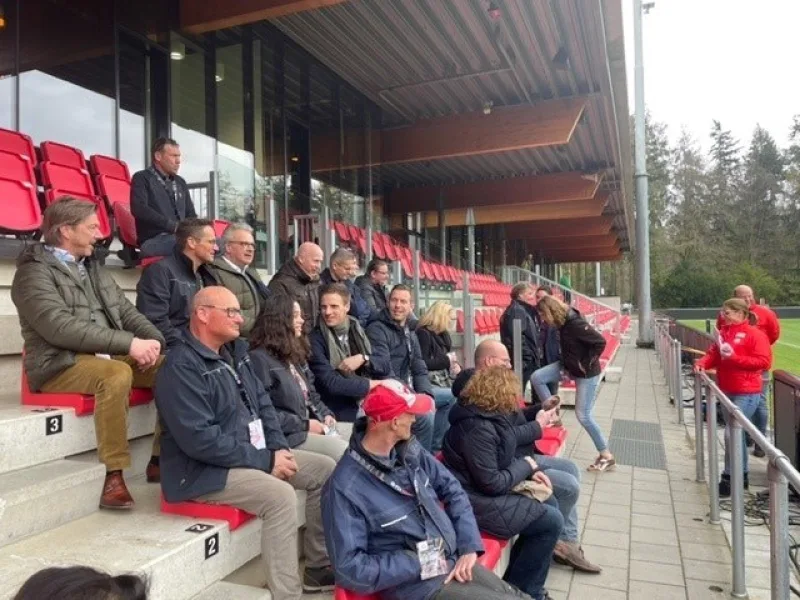 Groep bezoekers op rode tribune luistert naar uitleg langs voetbalveld