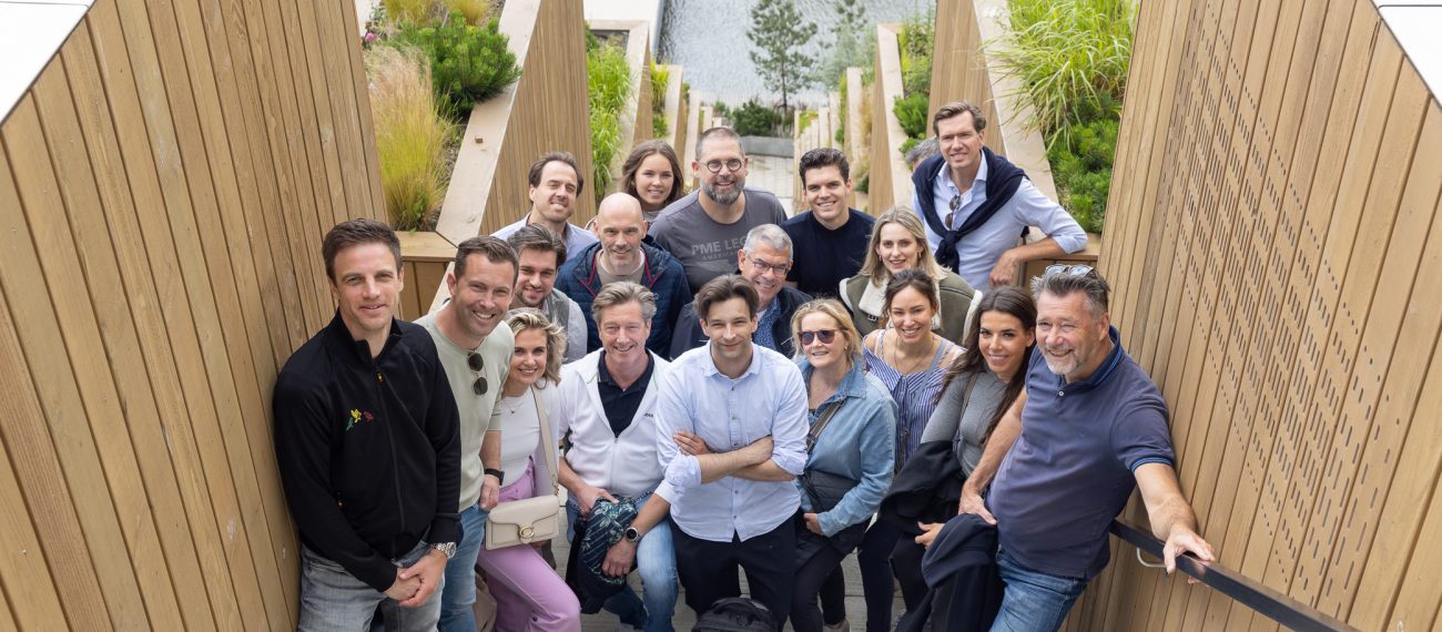 Groepsfoto van het team van CRA Vastgoed op een brede buitentrap met houten wanden en groen, met op de achtergrond een waterpartij. Het team staat dicht bij elkaar en kijkt lachend in de camera.