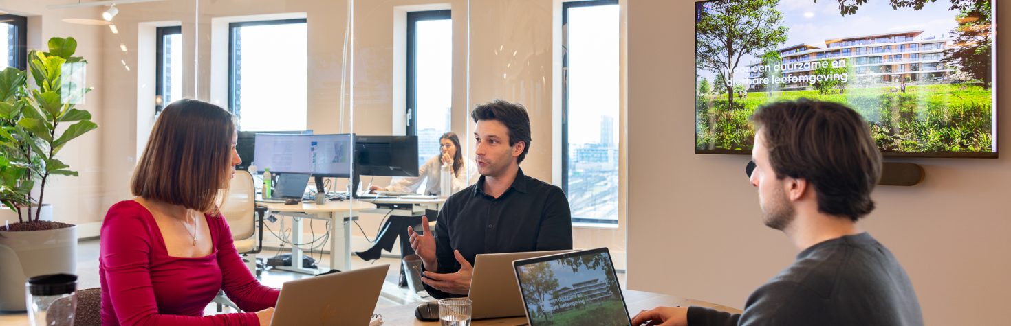 Drie collega’s zitten aan een vergadertafel met laptops tijdens een bespreking, met een presentatie op een scherm en een kantoorruimte op de achtergrond.