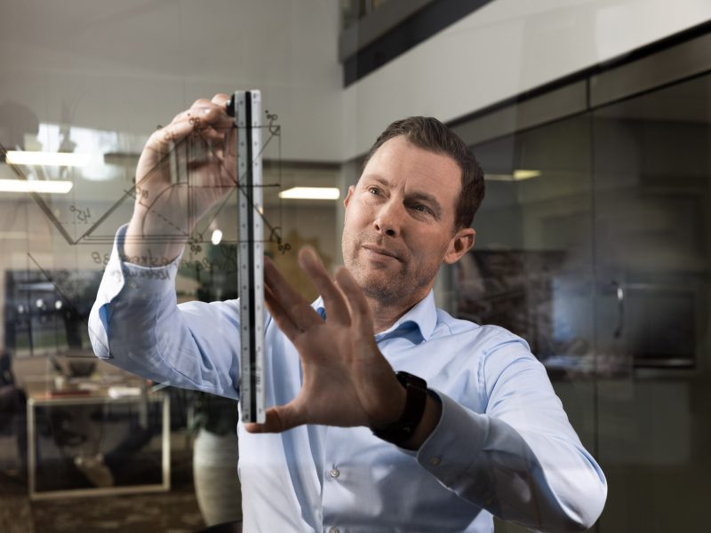 Man tekent met een liniaal op een glazen wand met technische schetsen in een kantooromgeving.