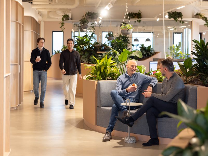 Twee mannen zitten pratend op een bankje in een modern kantoor met veel planten, terwijl twee andere mannen langslopen op de achtergrond