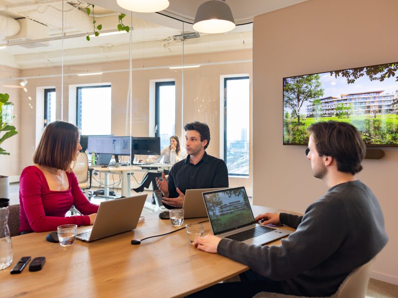 Drie collega’s zitten aan een vergadertafel met laptops tijdens een bespreking, met een presentatie op een scherm en een kantoorruimte op de achtergrond.