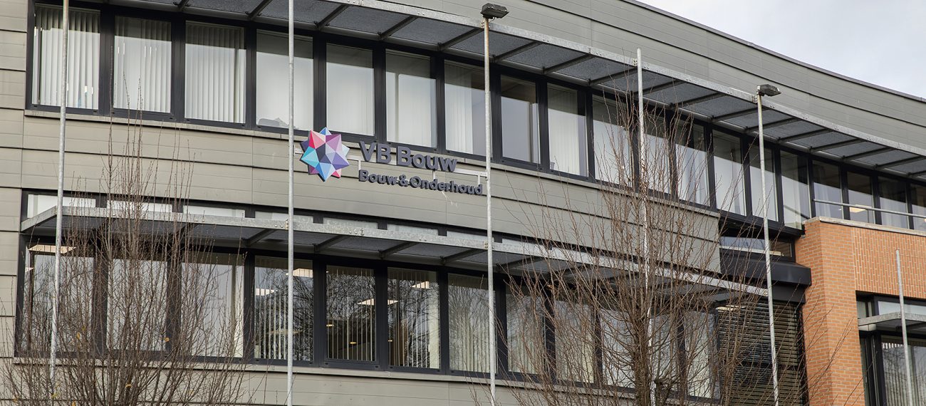 Moderne bedrijfspandgevel met groot logo en zwarte raamstroken, gezien vanaf straat met vlaggenmasten