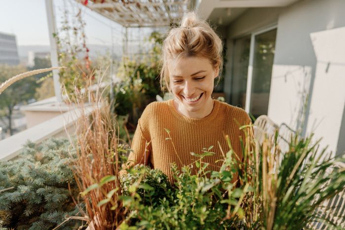 Vrolijke jonge vrouw tuiniert op haar zonnige balkon tussen weelderige planten en kruiden.