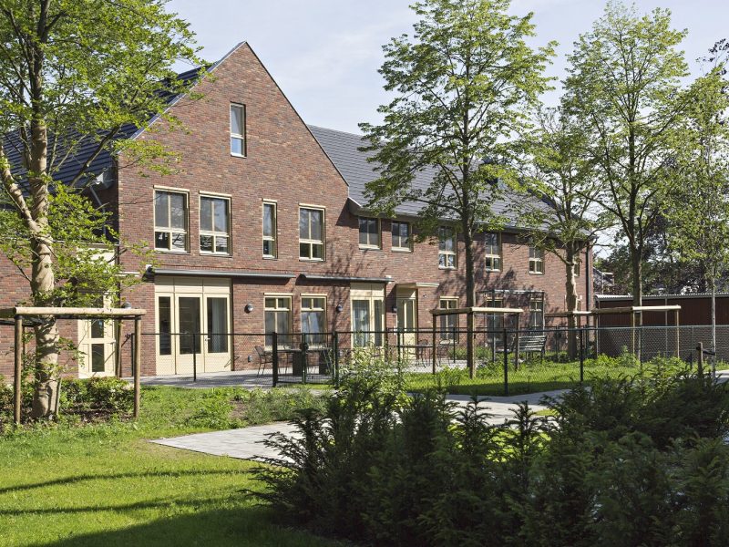 Rij moderne bakstenen woningen met tuin en groenvoorziening op een zonnige dag, met twee mensen die over een pad langs het gebouw lopen.