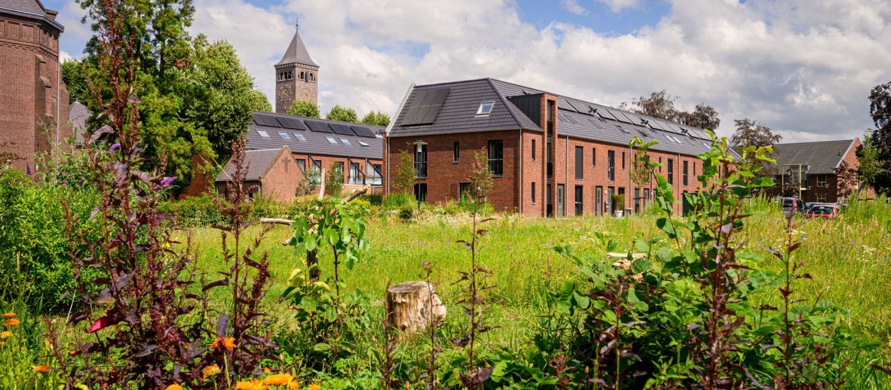 Gebiedsontwikkeling Kloostervelden in Sterksel met nieuwbouw rijwoningen in rood baksteen met zonnepanelen rond een groene, bloemrijke binnentuin en op de achtergrond een kloostertoren.