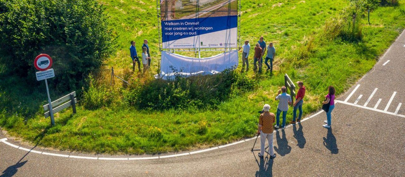 Groep mensen staat bij een groot bouwbord langs een landelijke weg.