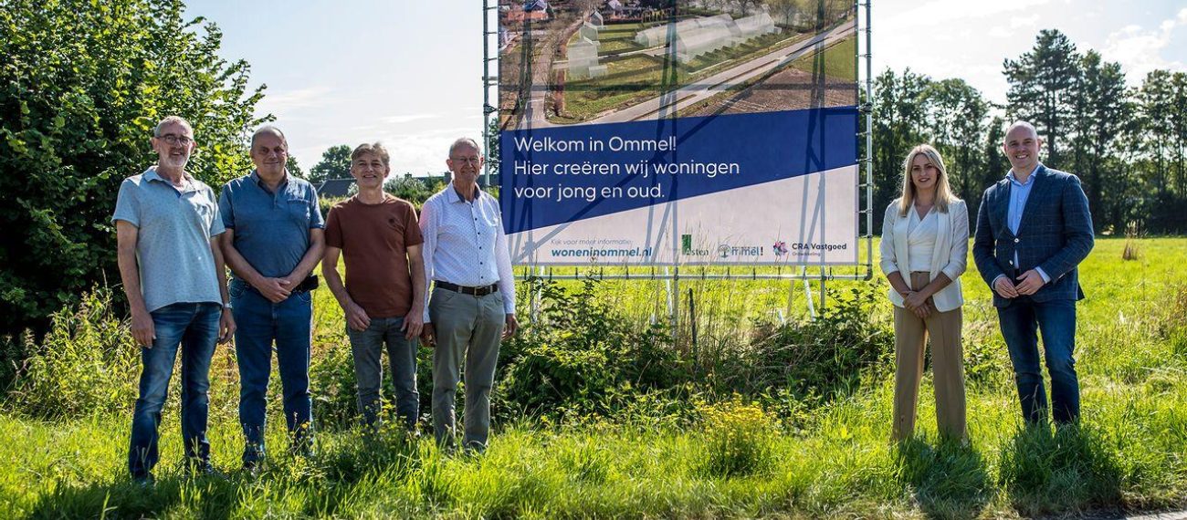 Groep mensen staat naast een groot bouwbord langs een groene weg.