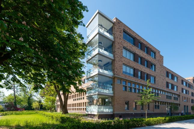 Modern woongebouw met bakstenen gevel en glazen balkons omringd door groen en bomen op een zonnige dag.
