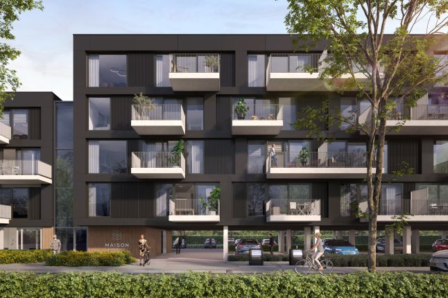 Close-up van Maison L’Europe in Helmond: modern donker appartementencomplex met uitstekende witte balkons en grote ramen, op kolommen met doorgang naar de parkeergarage; bomen, een fietser en voetgangers op de voorgrond.