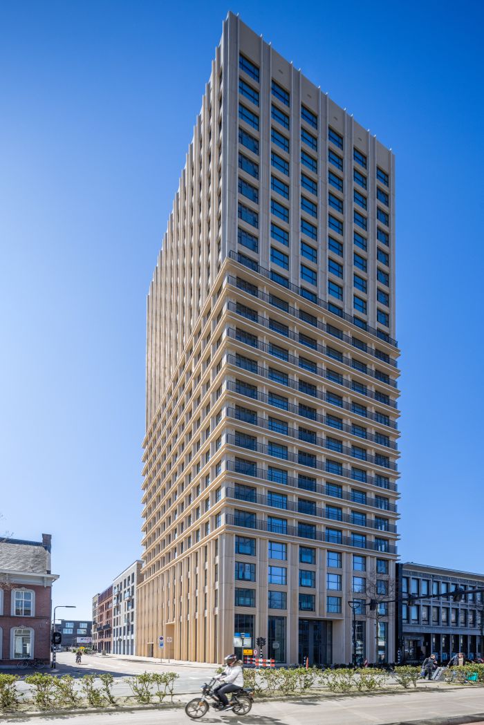 Ontwikkeling De Bankier Tilburg – volledig straatbeeld van de slanke woontoren met horizontale geveldetaillering en grote ramen, op de hoek bij het station onder een heldere blauwe lucht.