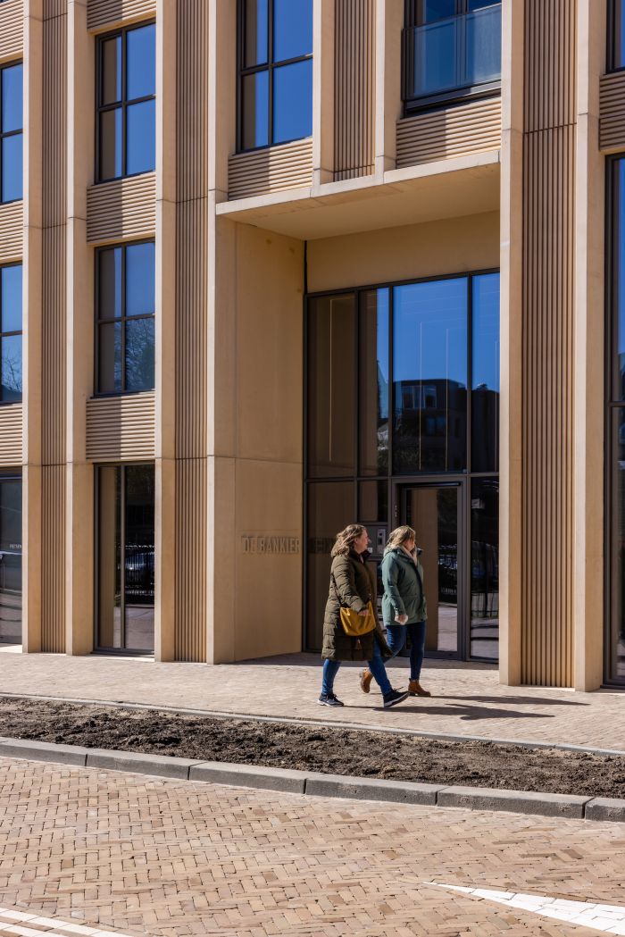Ontwikkeling De Bankier Tilburg – entreezone van de toren met hoge glazen pui en verticale geveldetaillering in zandkleurig beton, twee voorbijgangers op de stoep.