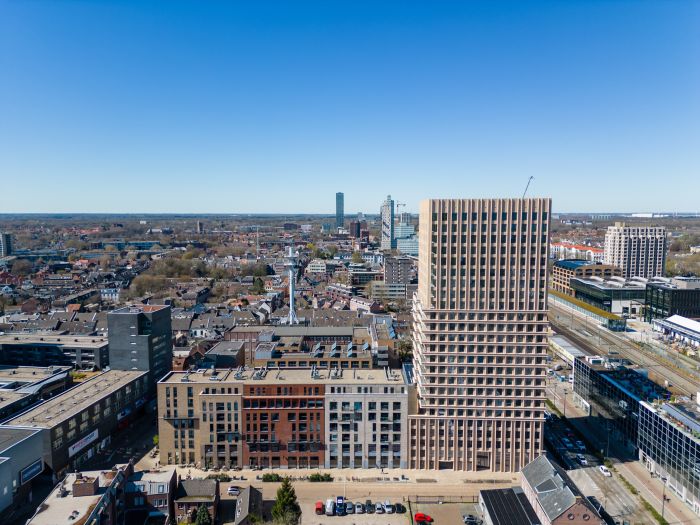 Ontwikkeling De Bankier Tilburg – luchtfoto met de nieuwe woontoren prominent in de skyline, naast het spoor en omliggende stedelijke bebouwing onder een heldere blauwe lucht.
