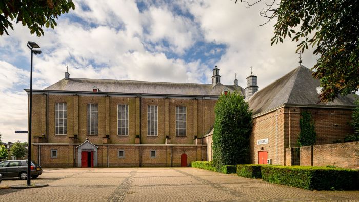 Transformatie Mariakerk Valkenswaard – zijaanzicht van de imposante bakstenen kerk met hoge verticale ramen en rode deuren, aangrenzend aan de koorkapel en kloosterzaal, met bestrate binnenplaats en strak gesnoeide hagen onder een deels bewolkte lucht.
