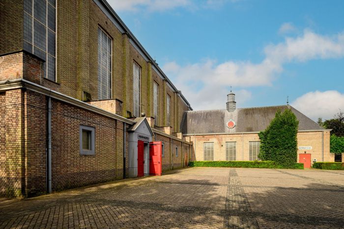 Mariakerk Valkenswaard binnenplaats – bakstenen kerkgevel met hoge glas-in-loodramen en open rode deuren, naast de kerkzaal met zadeldak en groen haagwerk onder een zonnige lucht.