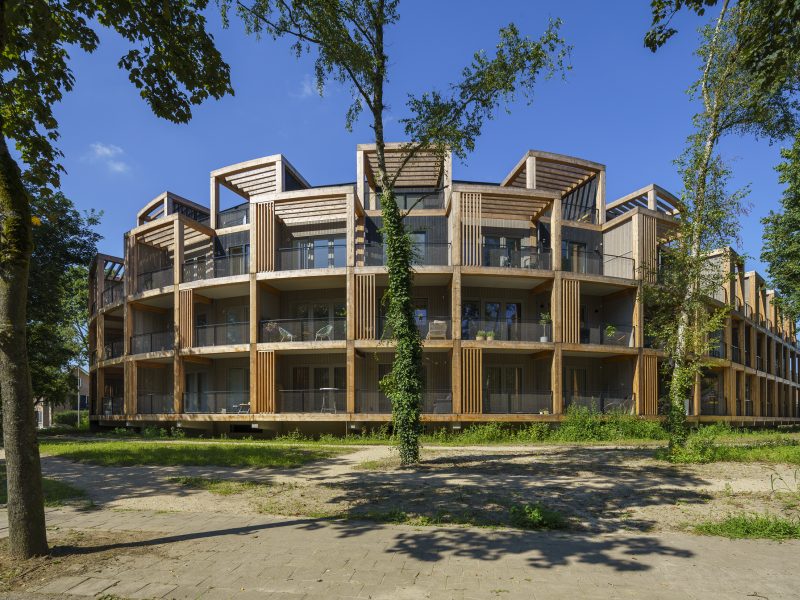 Rond woongebouw met meerdere verdiepingen en houten gevels, omringd door bomen en groen.