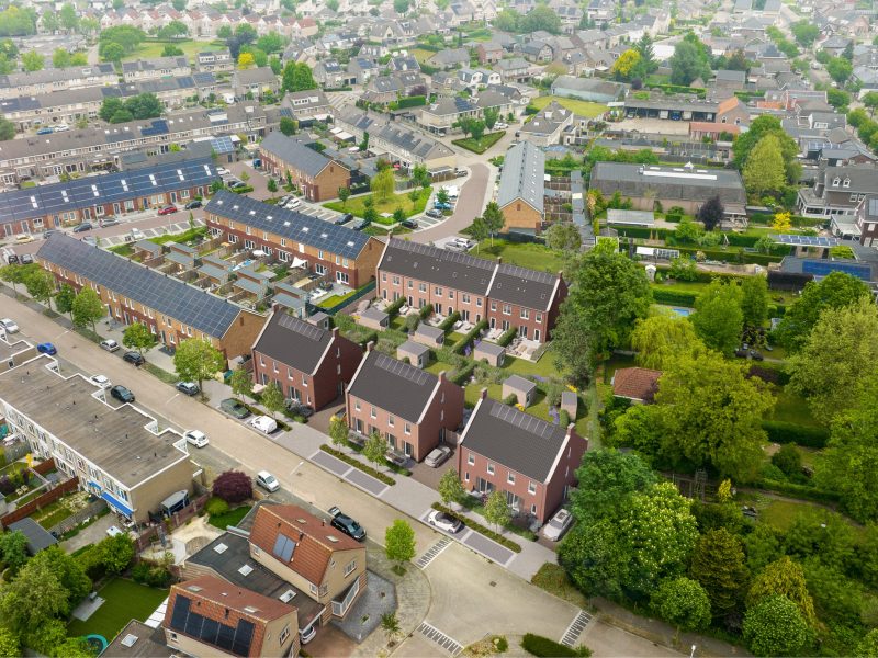 Luchtfoto van een woonwijk in Valkenswaard met in het midden een 3D-impressie van nieuwbouwproject Molensteen: drie bakstenen huizen met donkere zadeldaken en zonnepanelen, tuinen en parkeerplaatsen; op de achtergrond bestaande rijwoningen en veel groen.