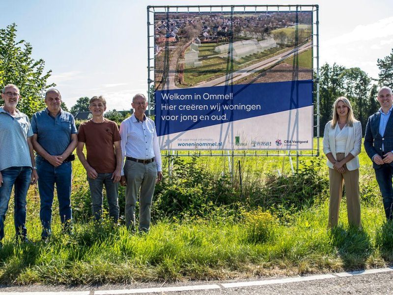 Groep mensen staat naast een groot bouwbord langs een groene weg.